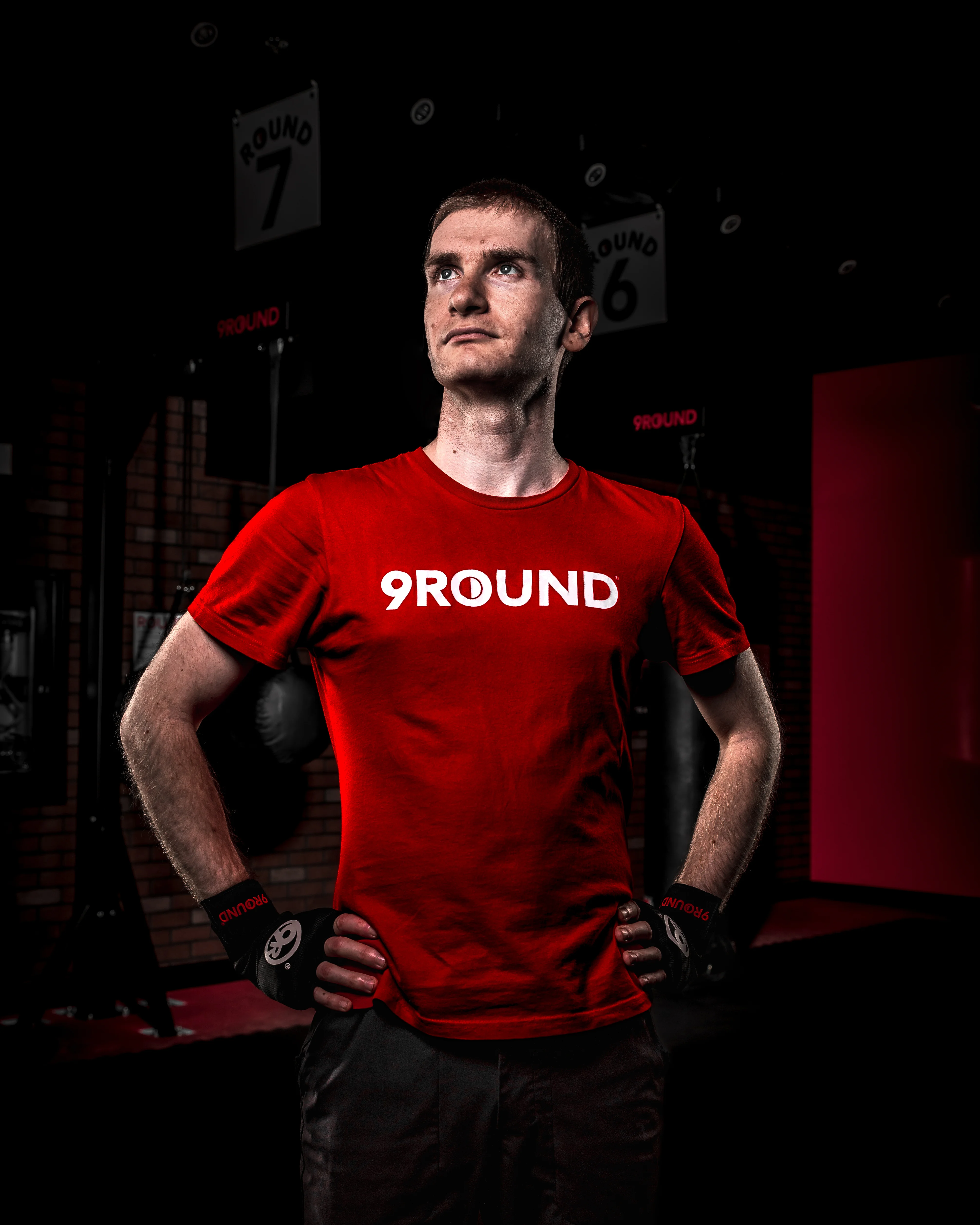 9Round fitness athlete in a red branded t-shirt standing confidently with hands on hips inside a boxing gym, dramatic low-key studio lighting with round number boards visible in background