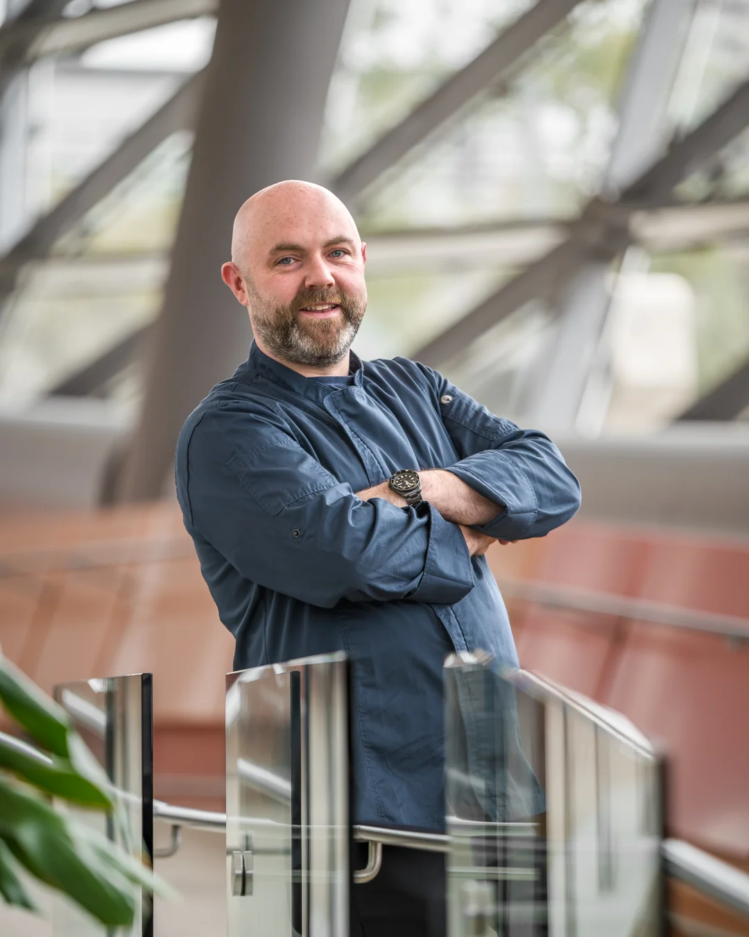 Male chef in a blue chef jacket smiling with arms crossed, leaning on a glass railing inside a bright modern atrium with a geometric lattice roof structure