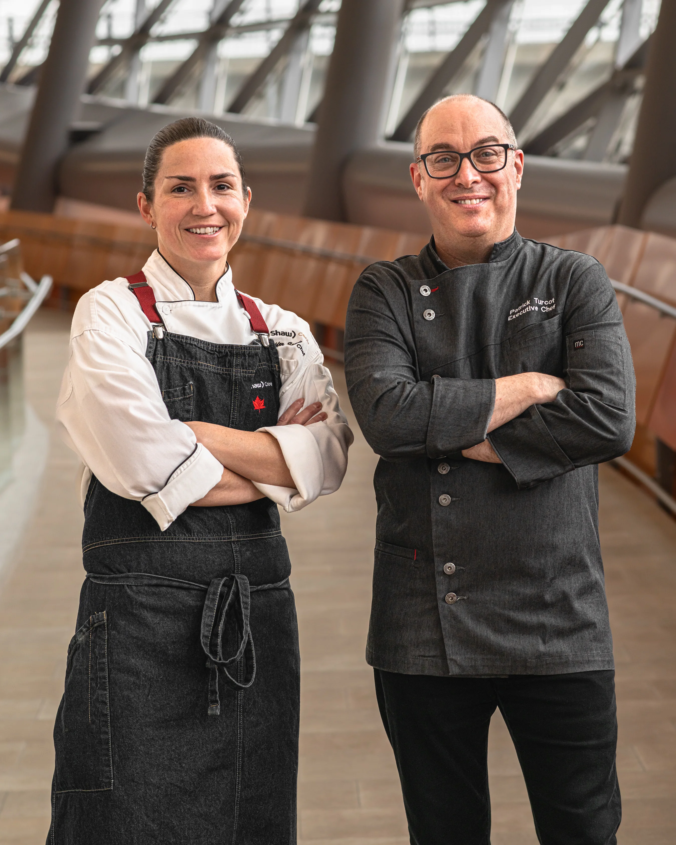 Female chef in a white jacket and male executive chef in a dark jacket standing confidently with arms crossed and smiling inside a modern architectural venue lobby