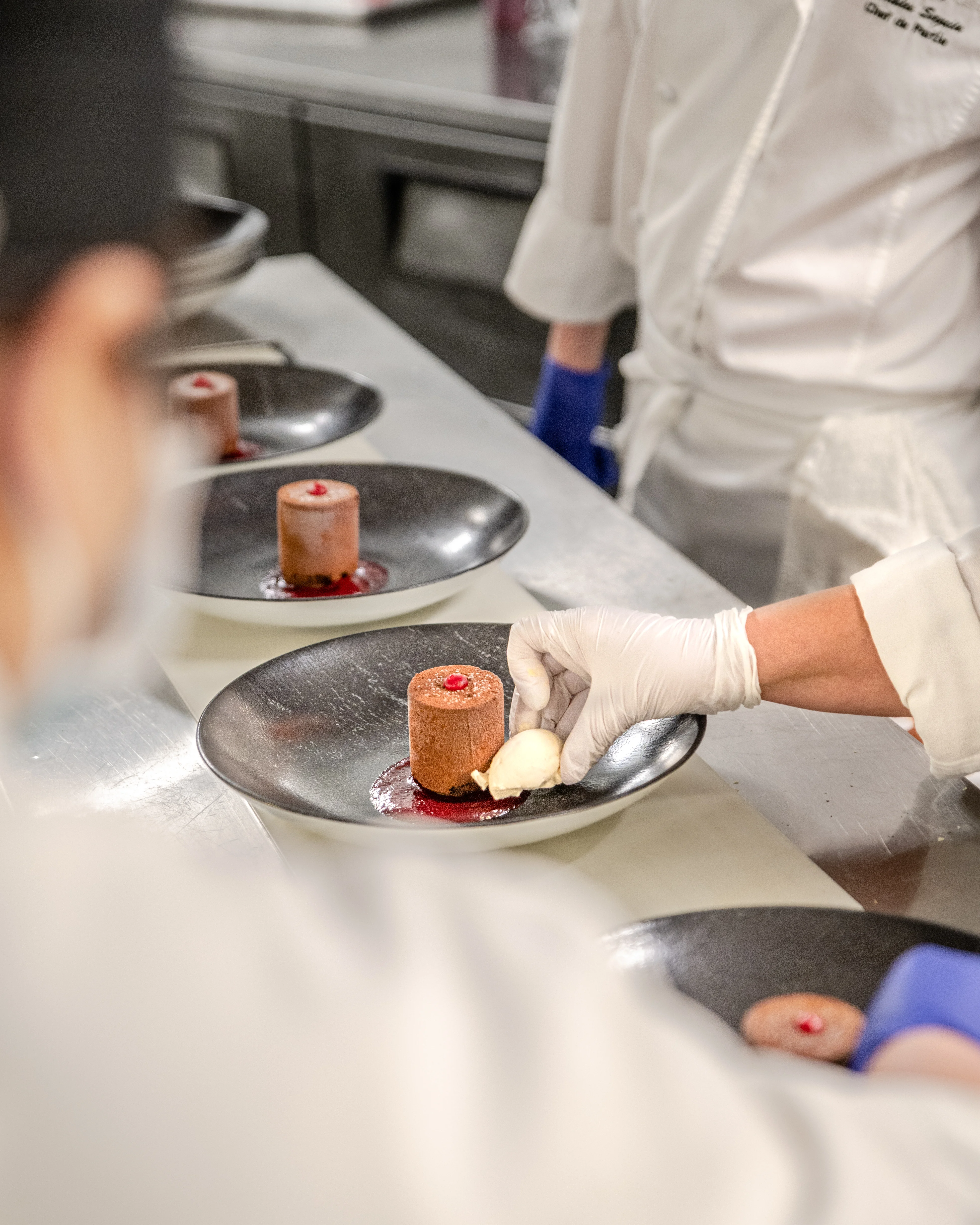 Kitchen team plating individual fine dining chocolate desserts in cylindrical molds with berry garnish on dark metal plates during a dinner service