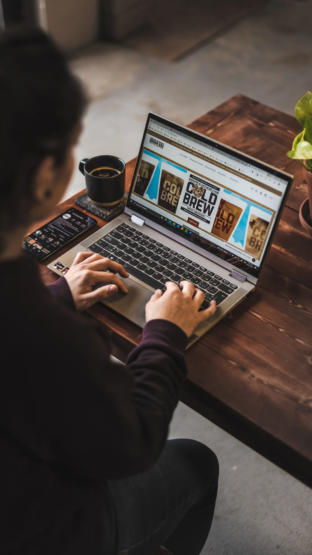 Person viewed from above typing on a laptop displaying a cold brew coffee brand website, with a black coffee mug and smartphone on a warm wooden desk
