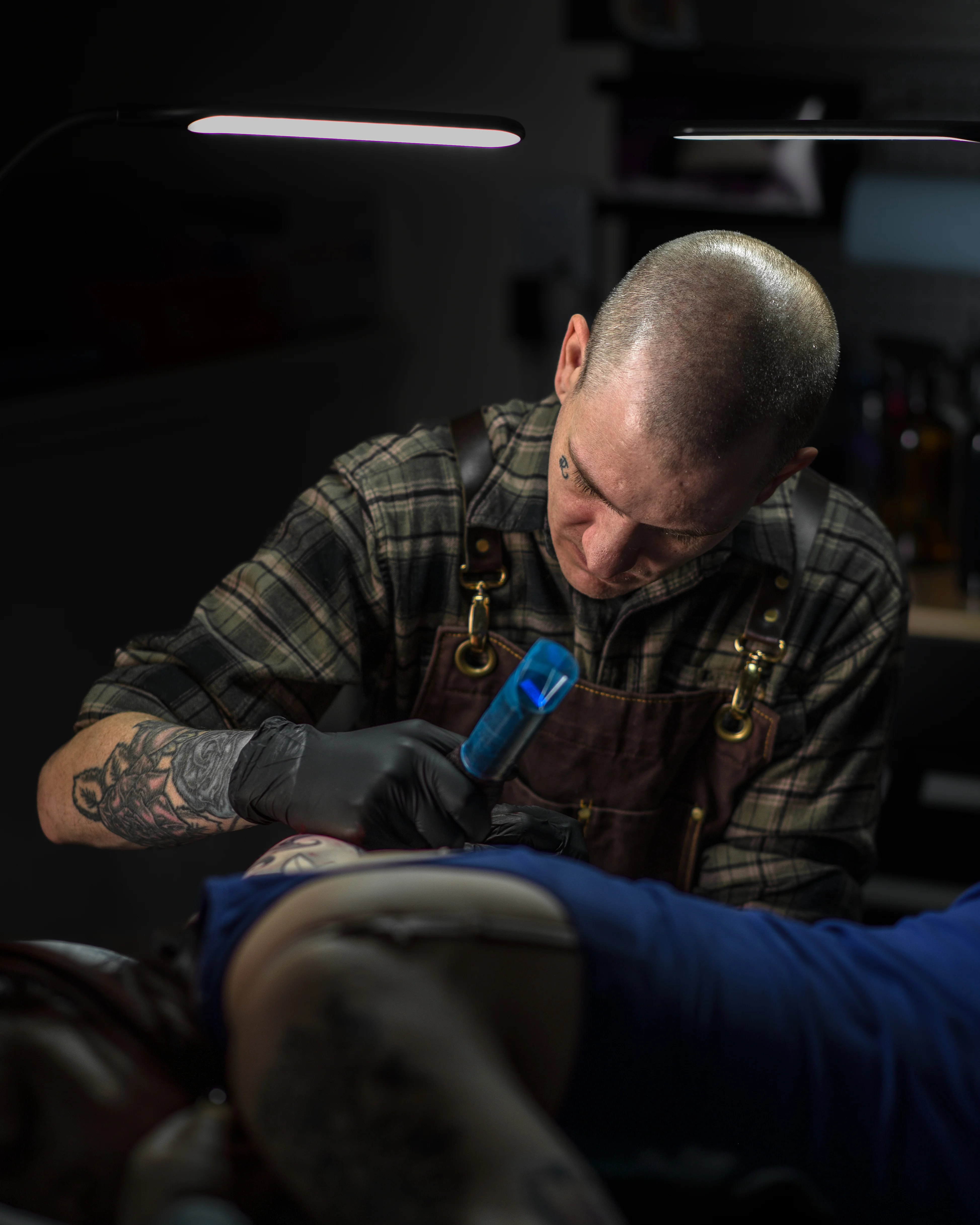 Tattoo artist with tattooed arms in a flannel shirt and leather apron concentrating while tattooing a client, illuminated by focused studio lights in a dark tattoo studio