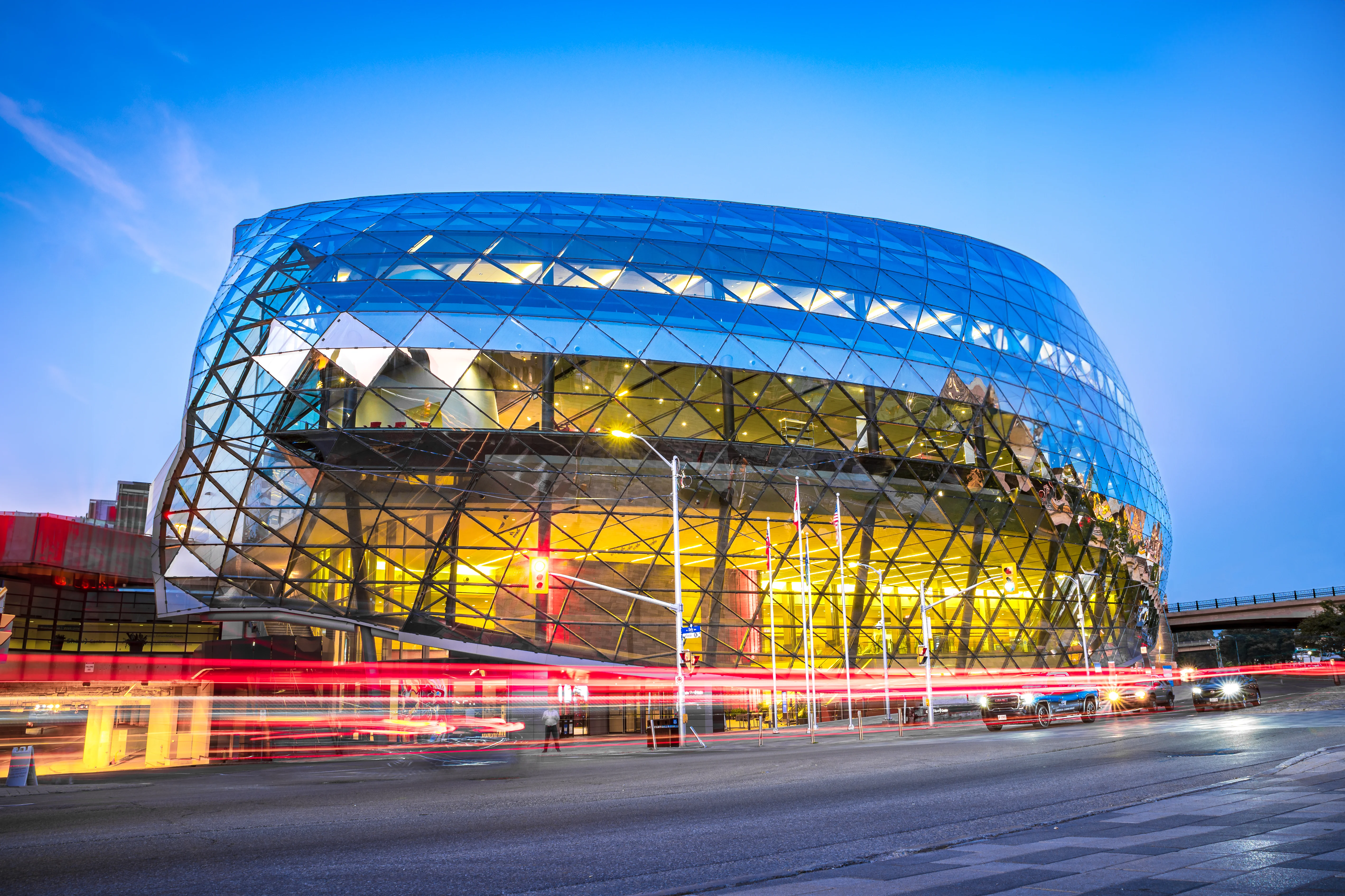 Modern arena building with a distinctive curved geometric glass and steel facade lit up with warm golden light, long-exposure light trails from passing traffic at dusk