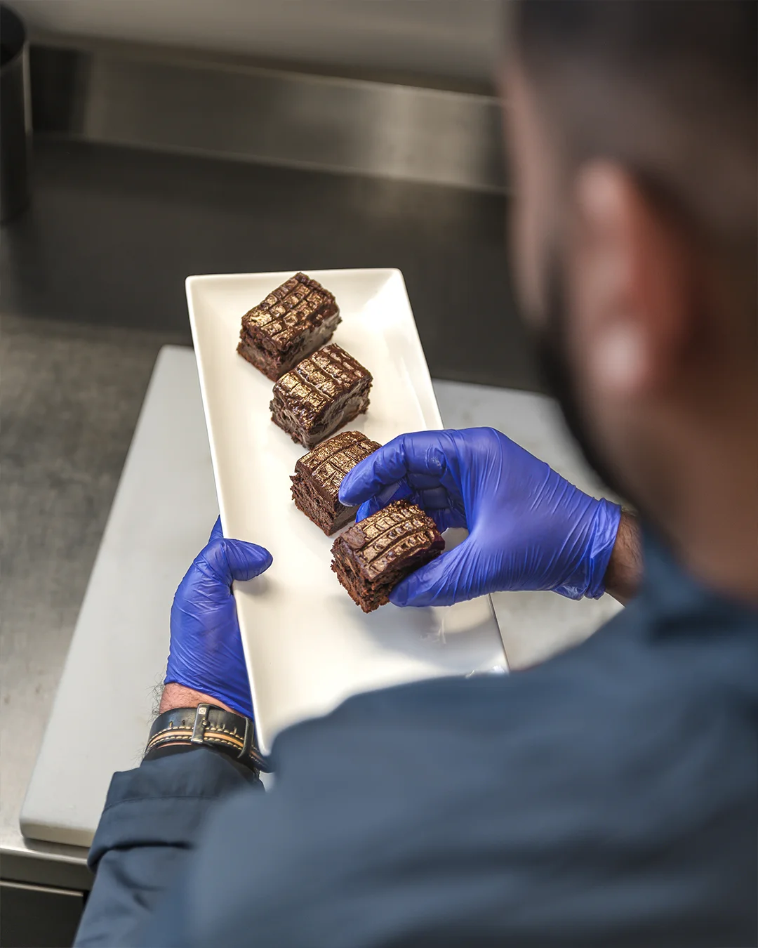 Female pastry chef seen from behind, carefully piping filling onto macaron shells lined up on a baking tray in a professional kitchen