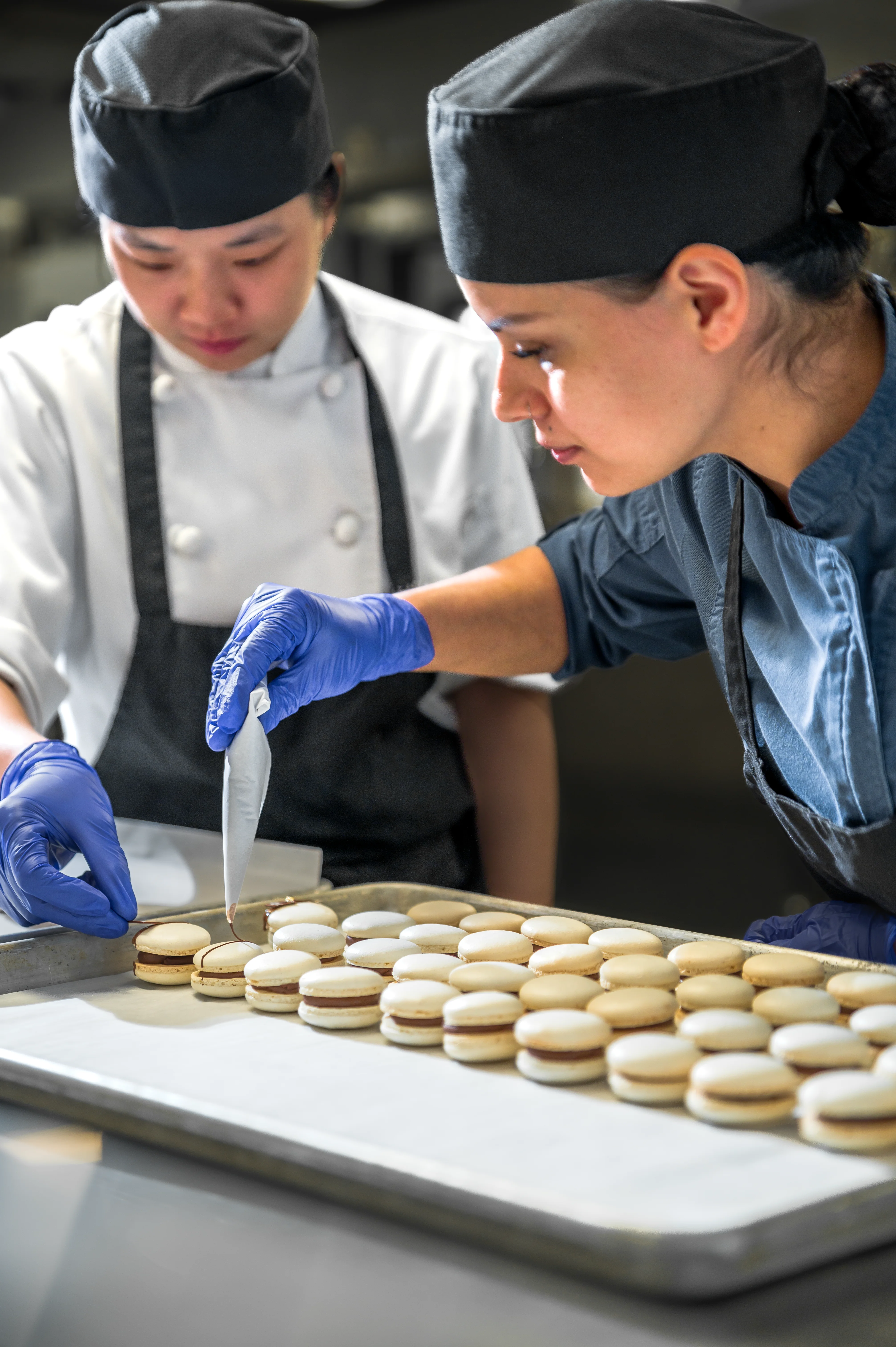 Two pastry chefs in black caps and blue gloves carefully filling assembled macaron shells on a baking tray in a professional commercial kitchen
