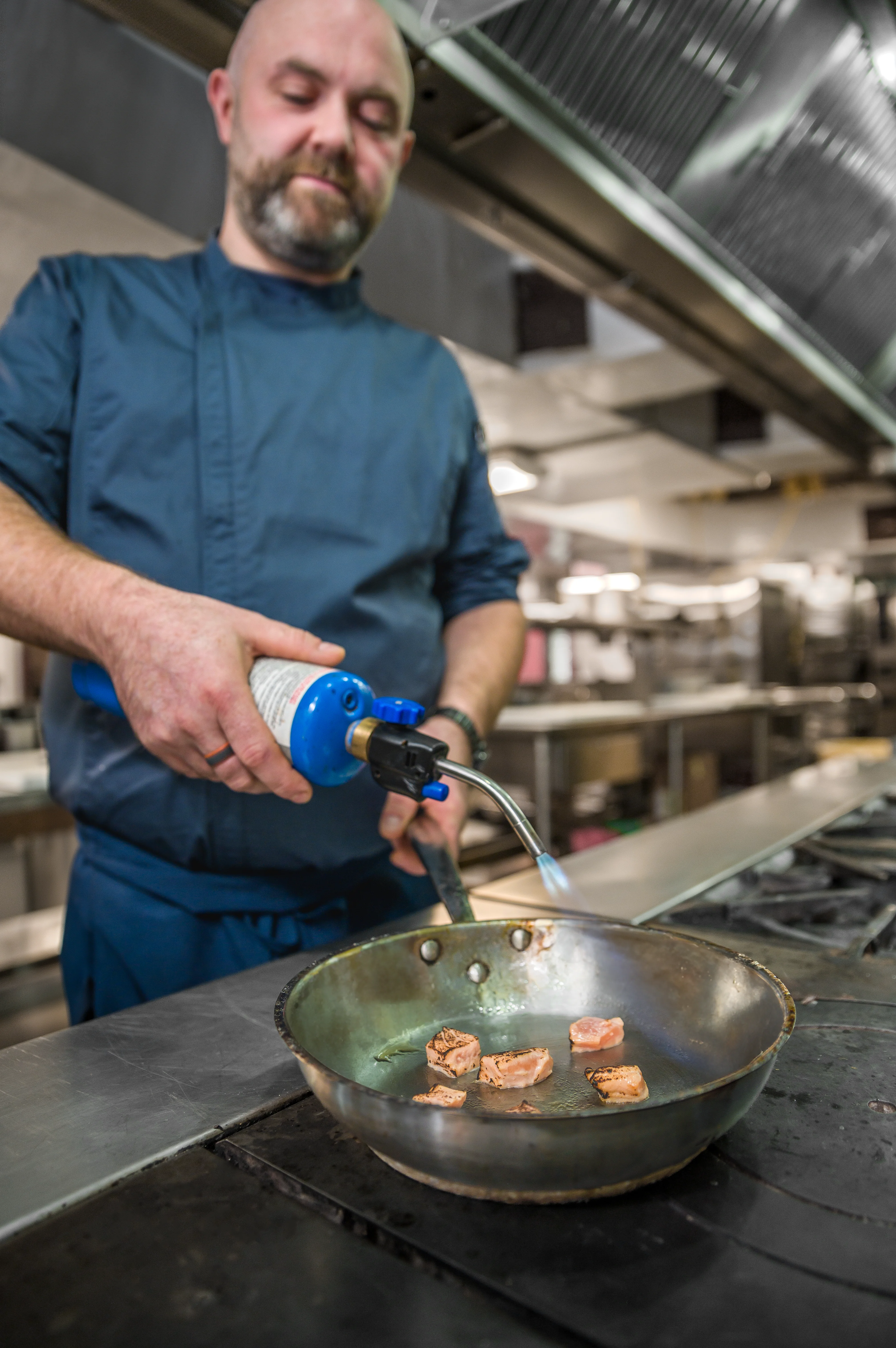 Male chef in a blue jacket using a culinary blowtorch to sear scallops in a cast iron pan on a commercial kitchen range