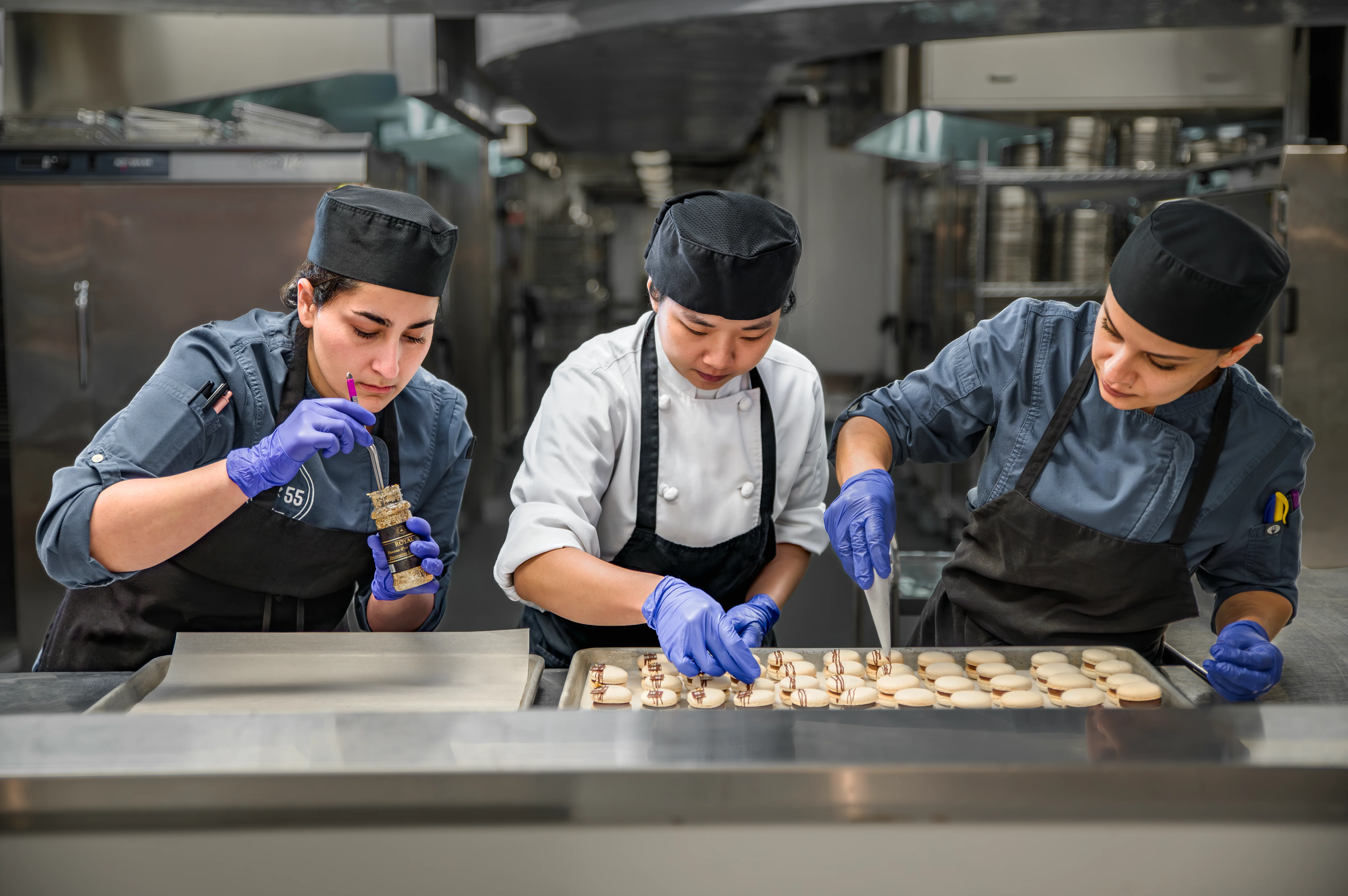 Three pastry chefs in a professional commercial kitchen collaborating to pipe and assemble small pastries on a baking tray beneath a stainless steel hood
