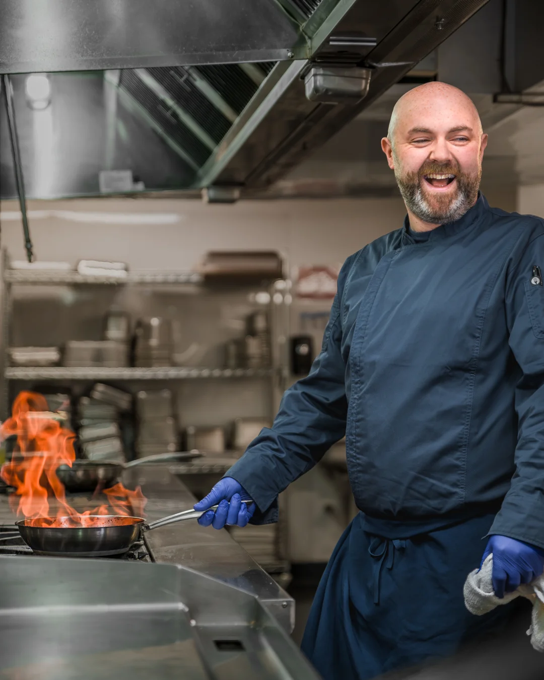 Male chef in a blue chef coat laughing joyfully while flambéing food in a pan over an open flame on a commercial kitchen range