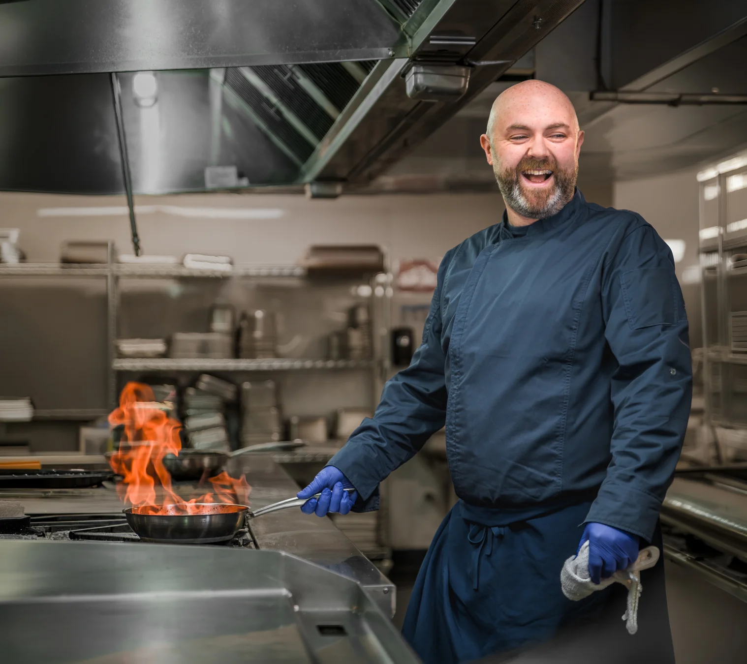 Male chef in a blue chef coat laughing while executing a flambé, wider shot showing more of the professional commercial kitchen environment