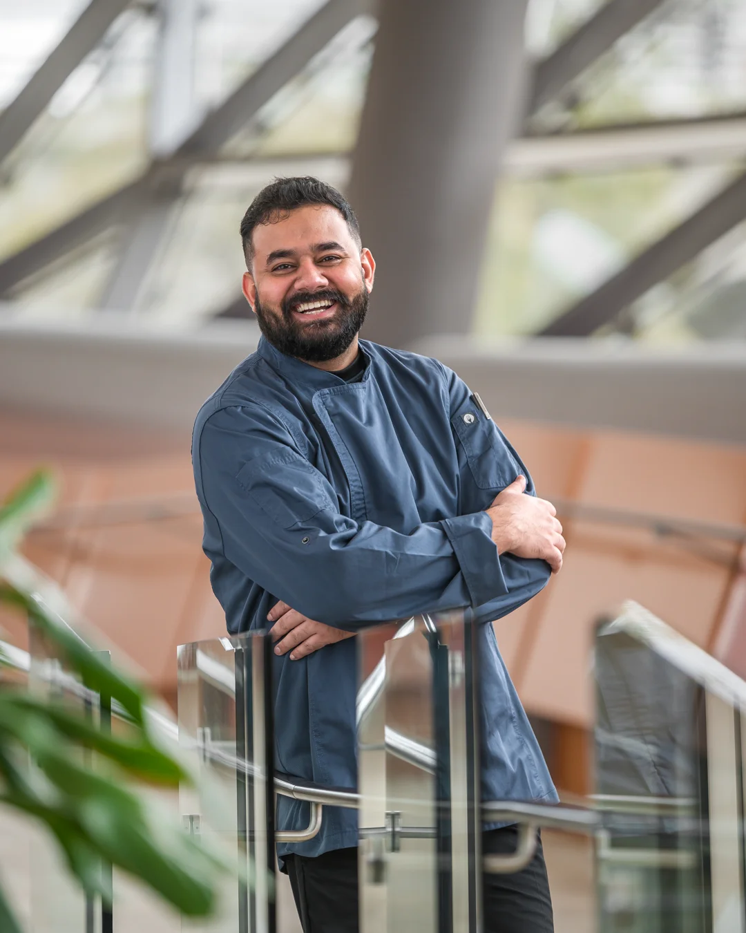 Smiling male chef in a blue chef jacket with arms crossed, leaning relaxed on a glass railing inside a sunlit modern atrium with a geometric steel roof structure