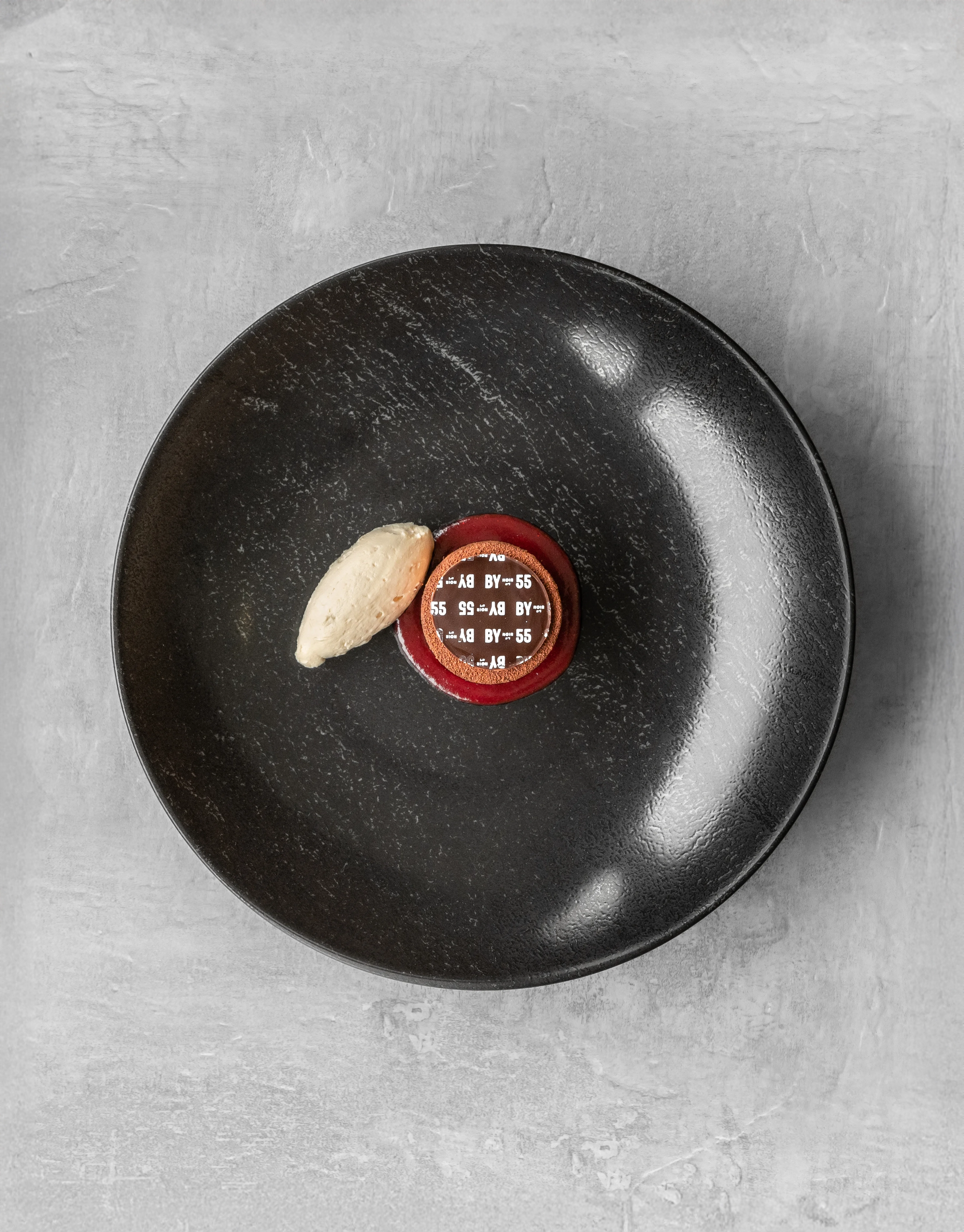 Overhead minimalist fine dining dessert — a small branded cylindrical chocolate mousse with a cream quenelle, presented on a dark matte black plate with a textured grey background