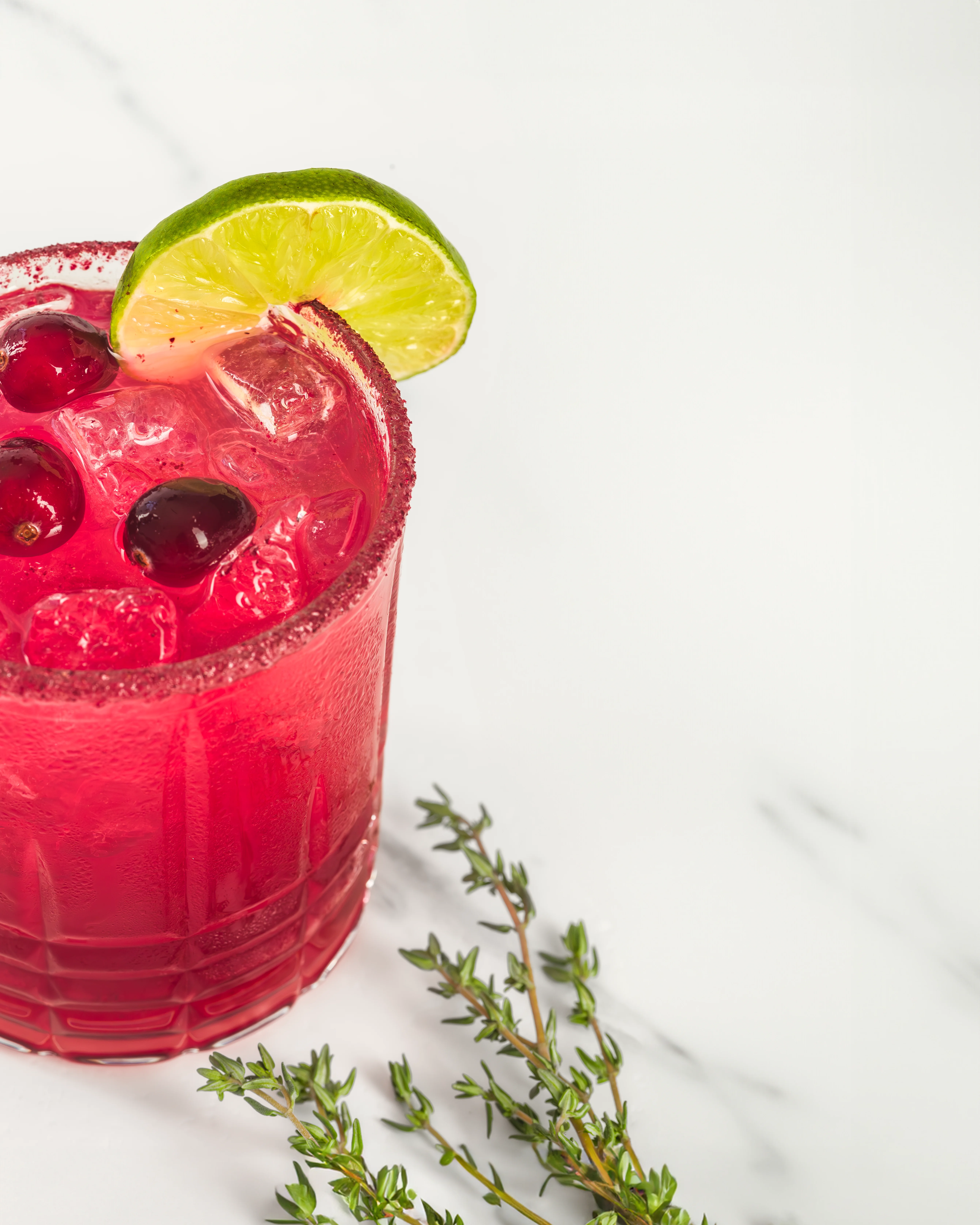 Vibrant cranberry cocktail in a textured rocks glass with a dark sugar rim, floating cherries, a lime wheel, and fresh thyme, displayed on a white marble surface