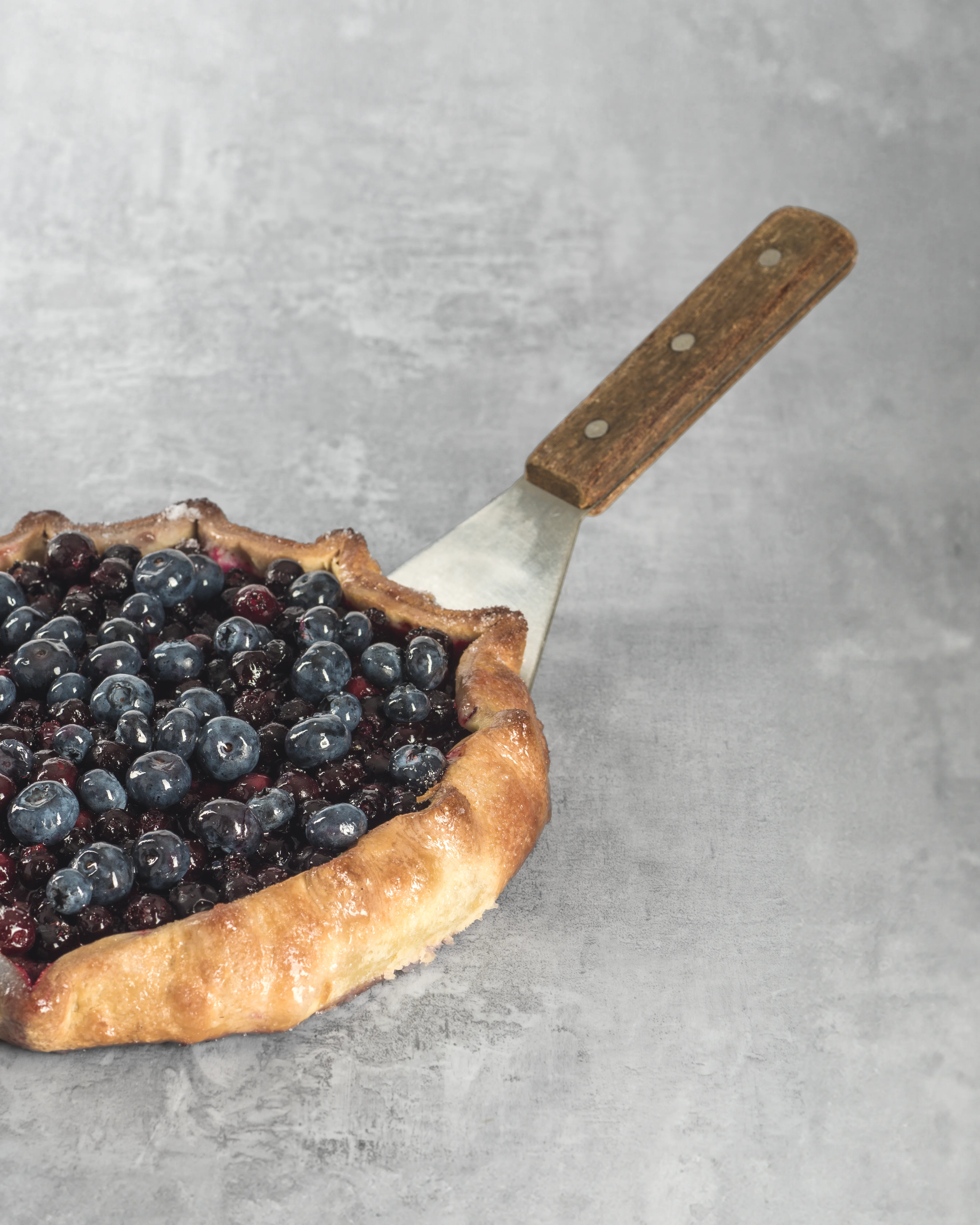 Side-angle view of a blueberry galette with a golden flaky pastry crust on a vintage spatula, grey concrete surface
