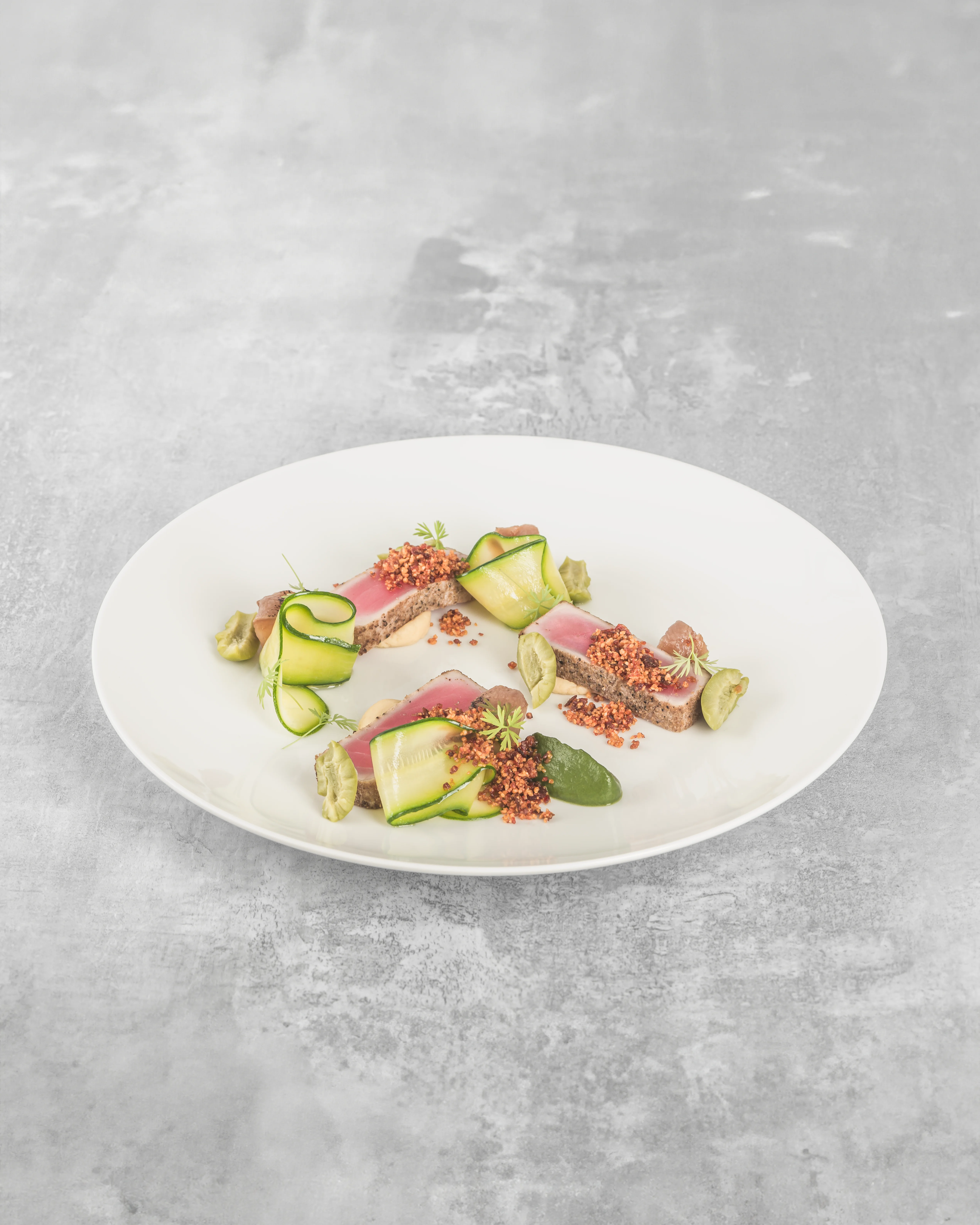 Fine dining seared tuna tataki with ribbons of cucumber, edamame, microgreens, and orange buckwheat crumble on a white plate against a grey concrete surface