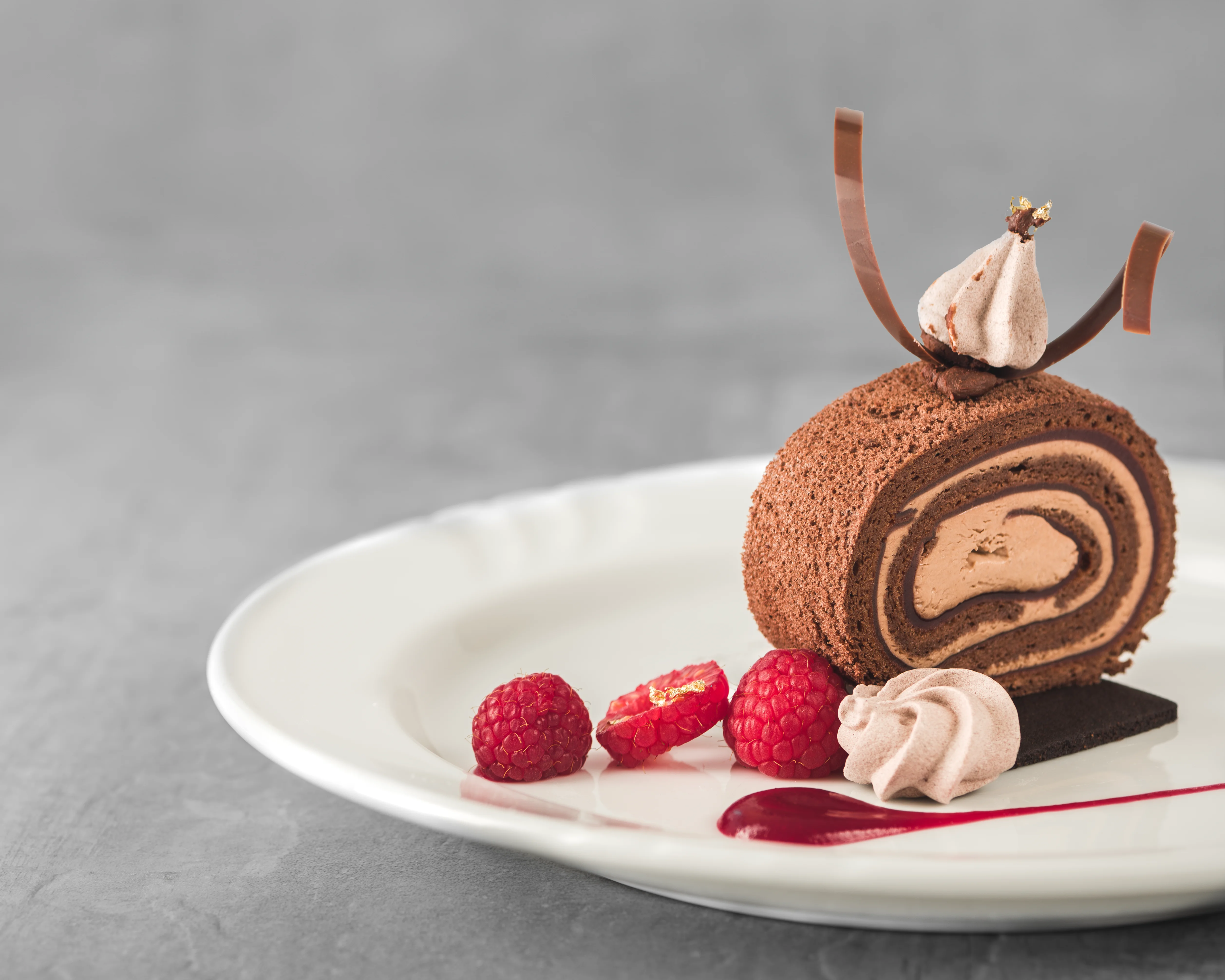 Chocolate roulade dessert plated with piped mocha cream, gold-dusted raspberries, chocolate curls, and a raspberry coulis drizzle on a white plate, grey background