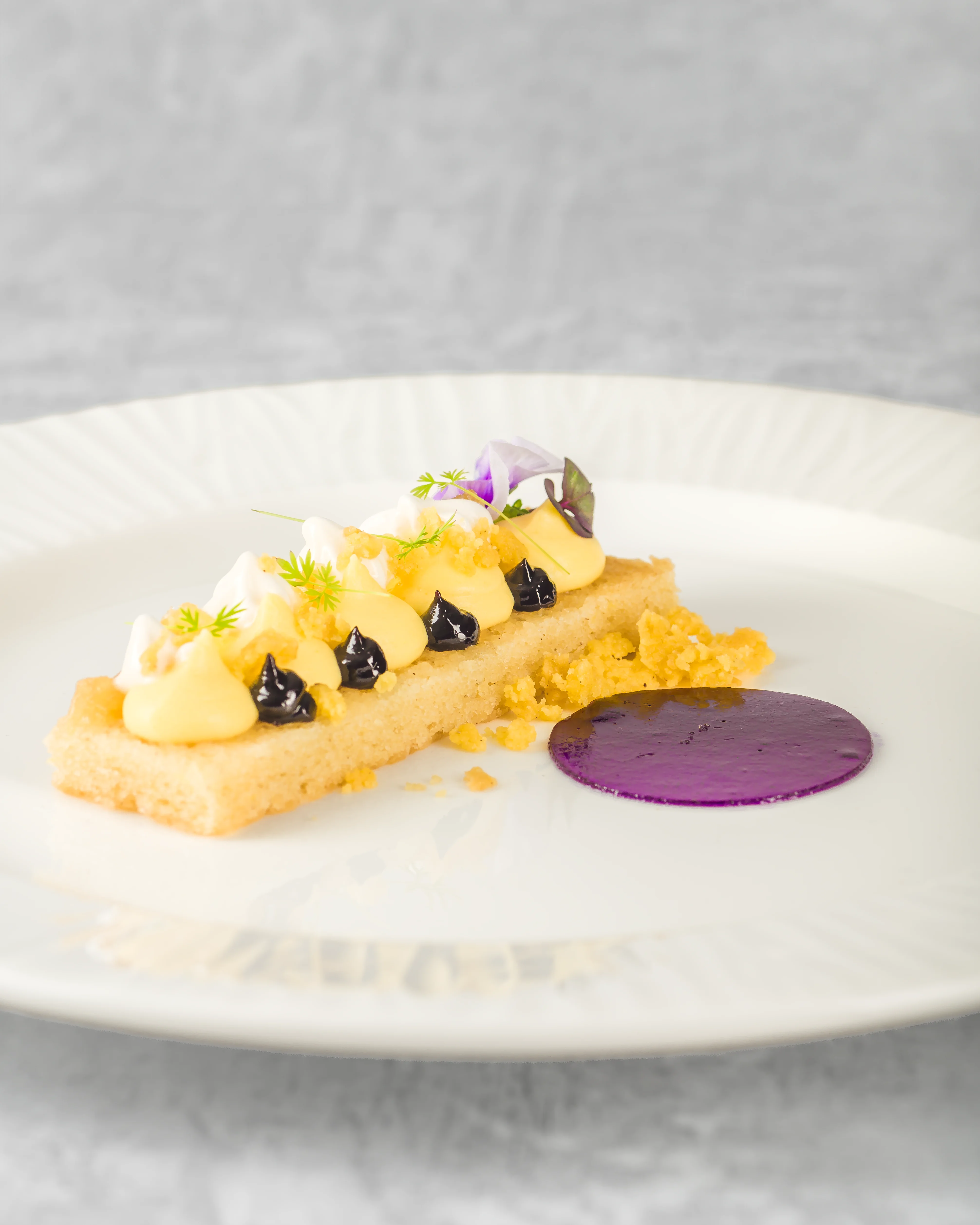 Elegant fine dining lemon tart with blueberry gel dots, edible flowers, custard quenelles, and crumble on a rectangular pastry base, served on a white ribbed plate