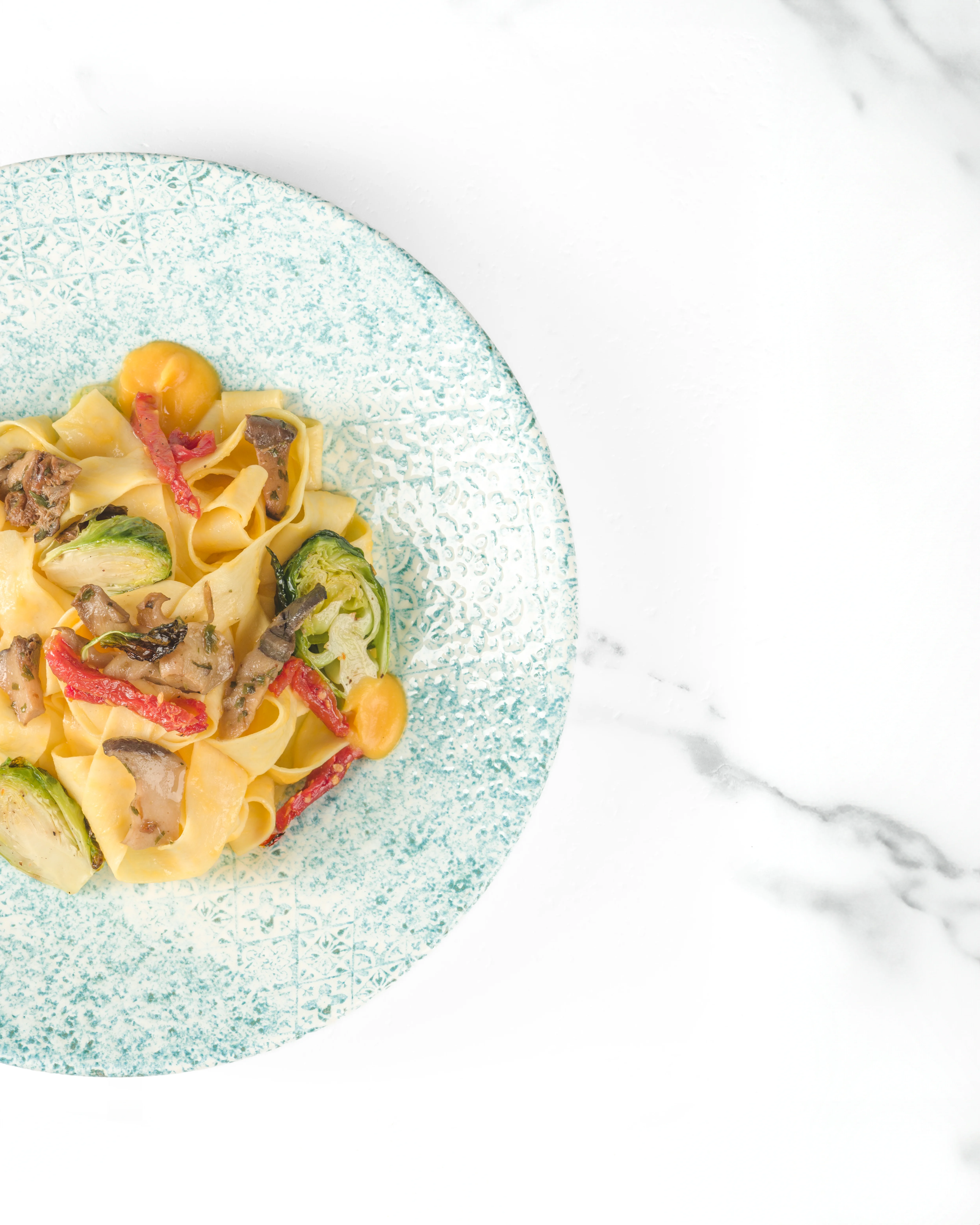 Overhead view of fresh pappardelle pasta with roasted mushrooms, zucchini, red pepper strips, and cherry tomatoes in a light blue speckled bowl on a white marble surface