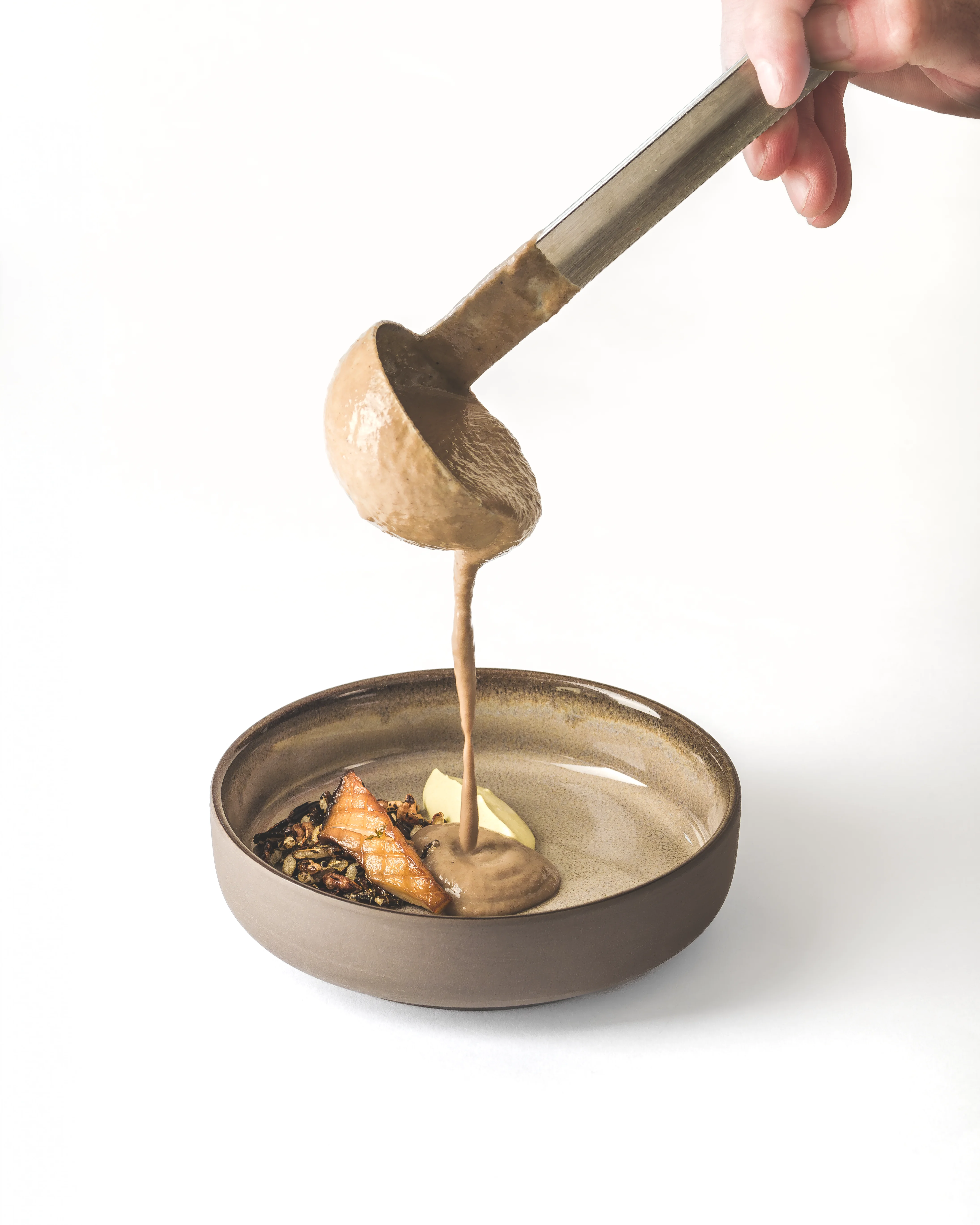 Dramatic action pour of a rich brown sauce from a ladle into a fine dining bowl containing seared salmon, wild rice, and butter, white background