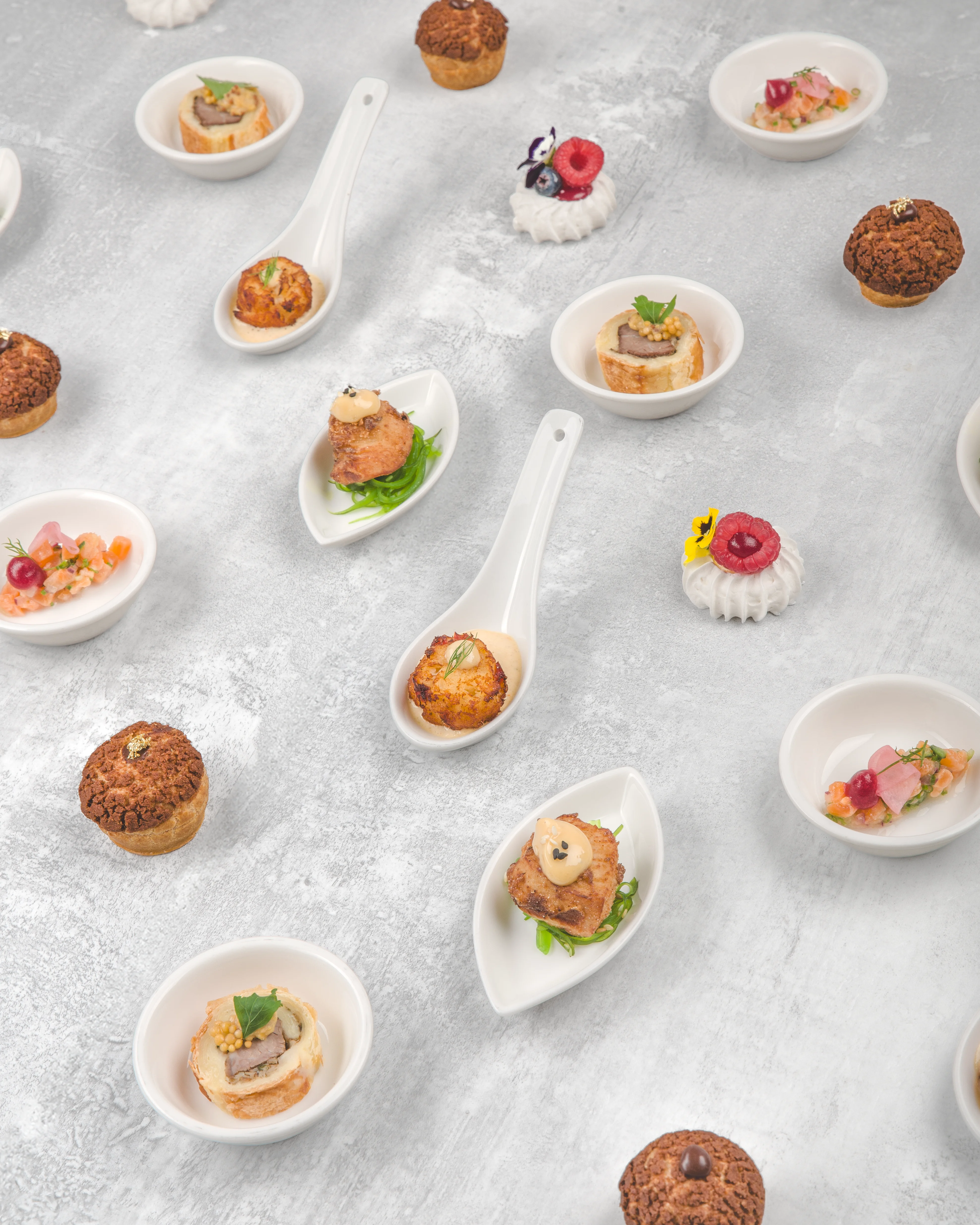 Overhead flat-lay of assorted gourmet canapés and hors d'oeuvres arranged on ceramic tasting spoons and small dishes across a grey concrete surface