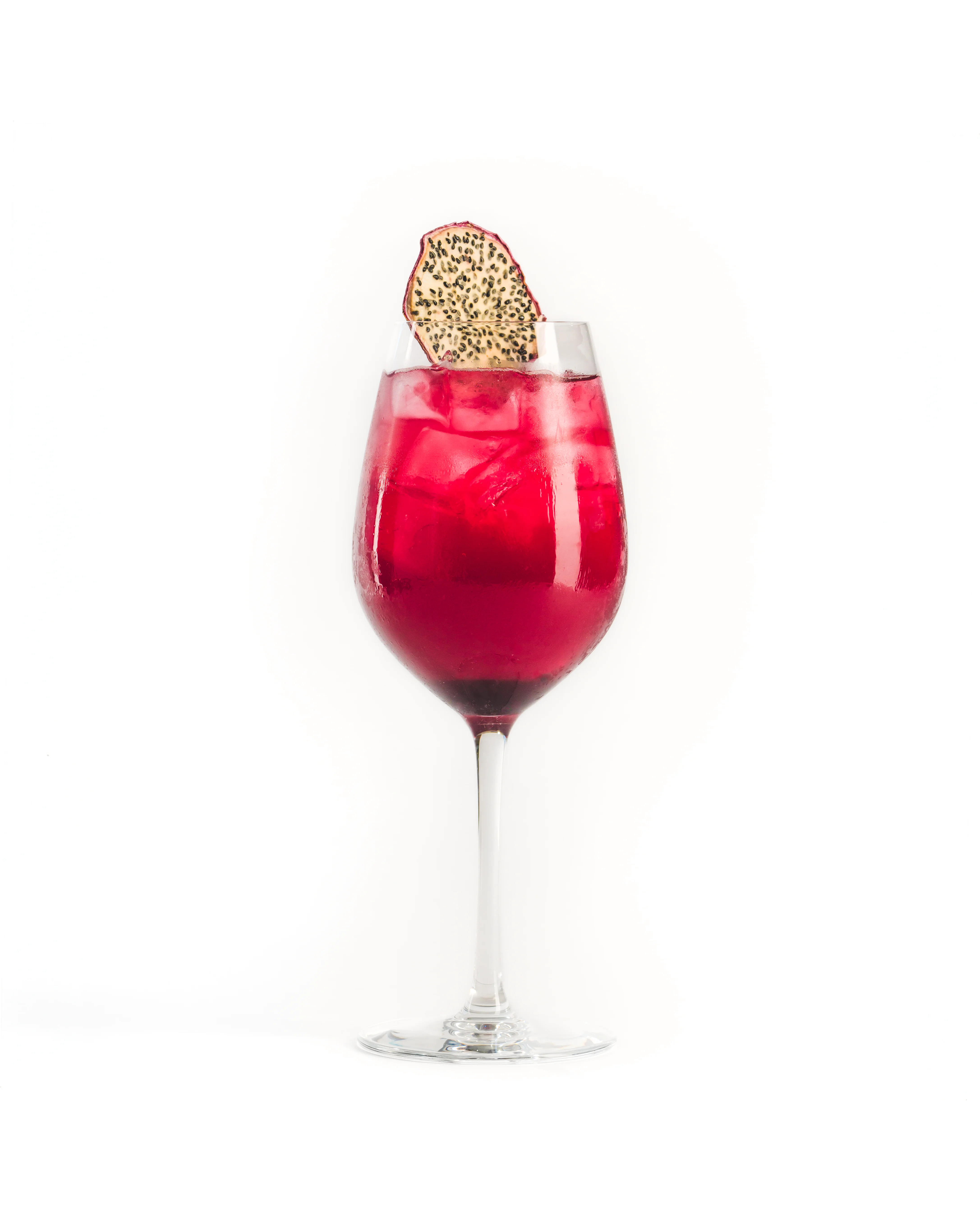 Deep red hibiscus cocktail in a large wine glass garnished with a dried dragon fruit wheel, photographed against a clean white background
