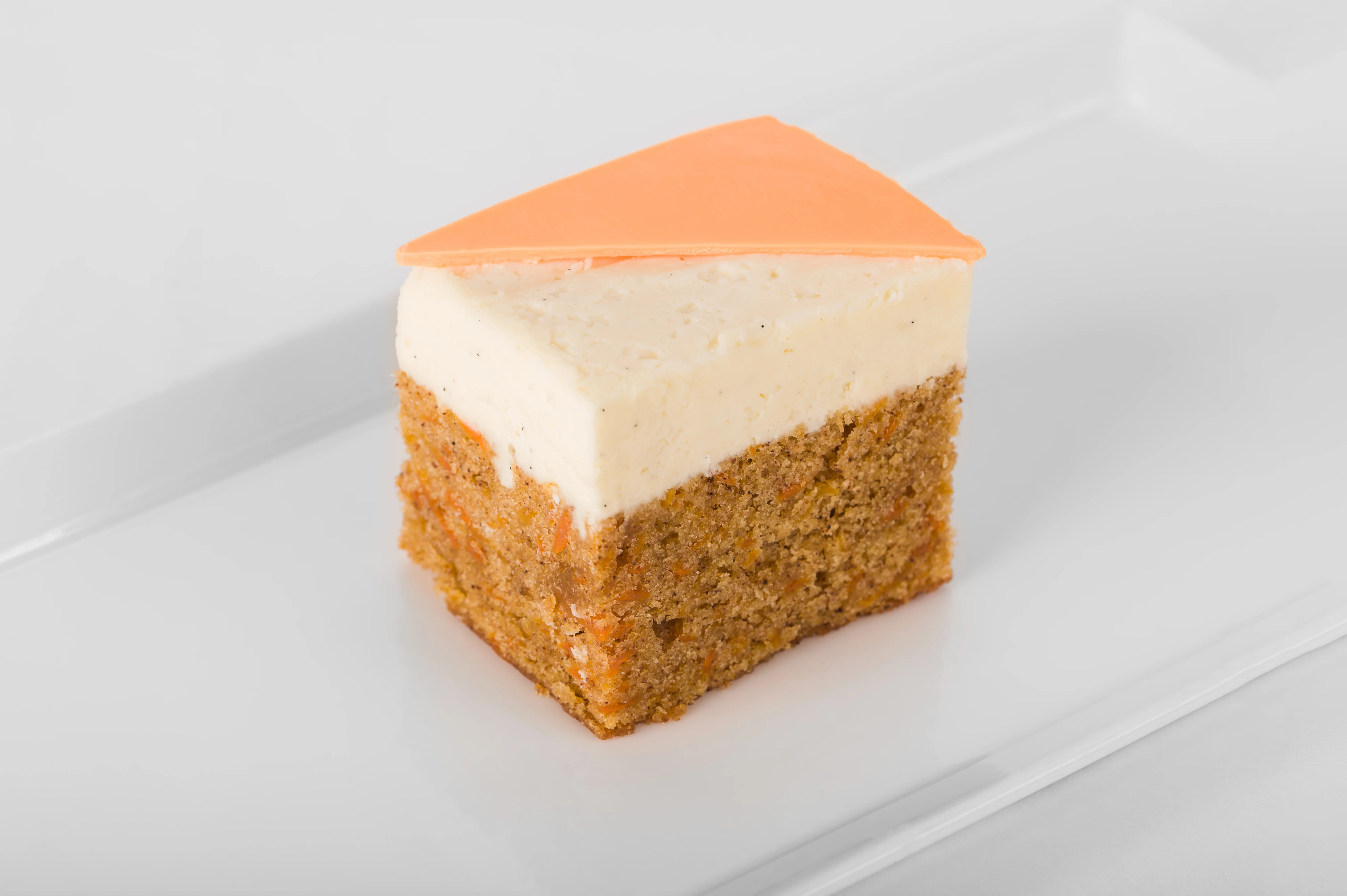 Carrot cake slice with cream cheese frosting and a glossy orange mirror glaze triangle, plated on a white rectangular plate, white background