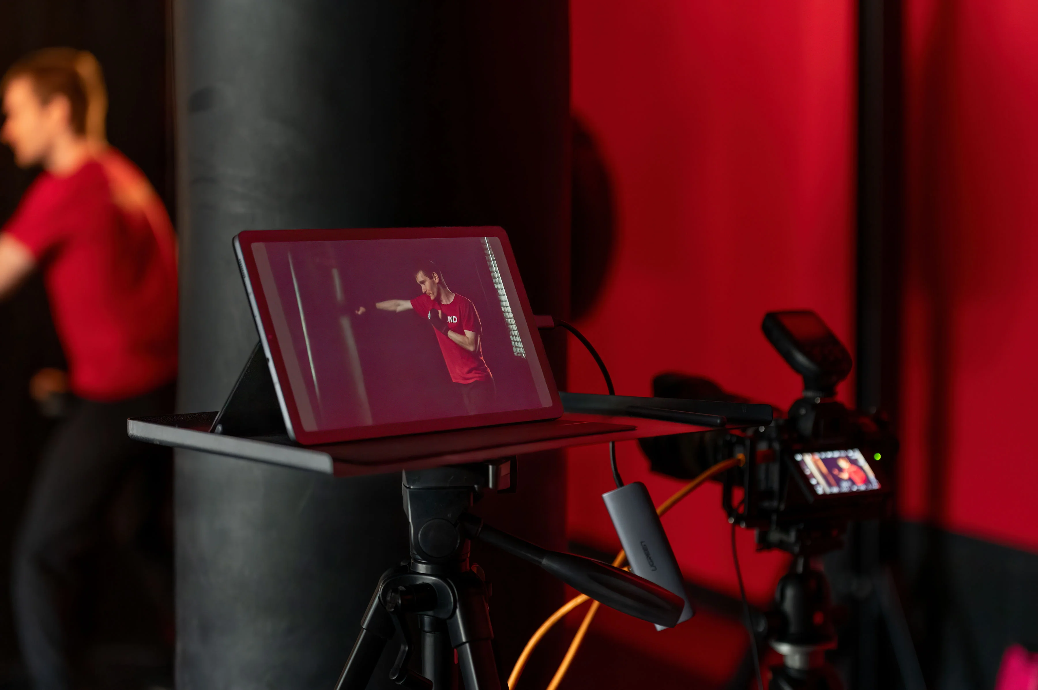 Behind-the-scenes view of a commercial photography studio shoot — a tablet on a stand showing an in-progress portrait of a 9Round athlete in red, with camera equipment and a subject visible in the soft background
